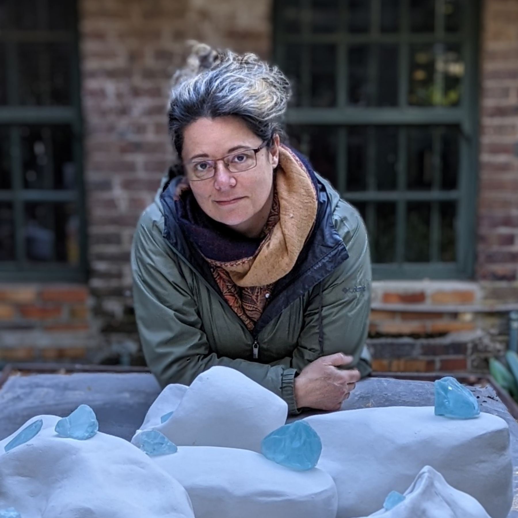 Lysanne Larose. A white woman leans forward, with her elbows on a table that displays white and ice blue sculptures. She is outside. She wears glasses, a green coat and a scarf. Behind her, a brick wall with windows.